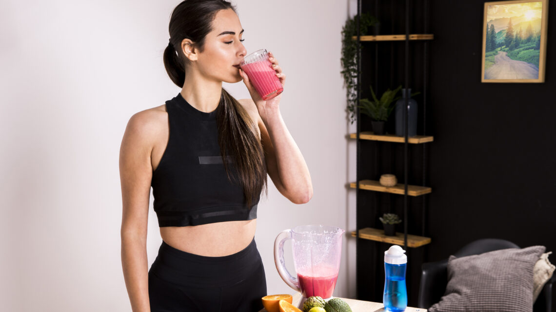 A fit woman wearing a black workout outfit drinking a fresh fruit smoothie beside a table of colorful ingredients, highlighting post-workout recovery habits inspired by a 24/7 gym in Greater Noida West.