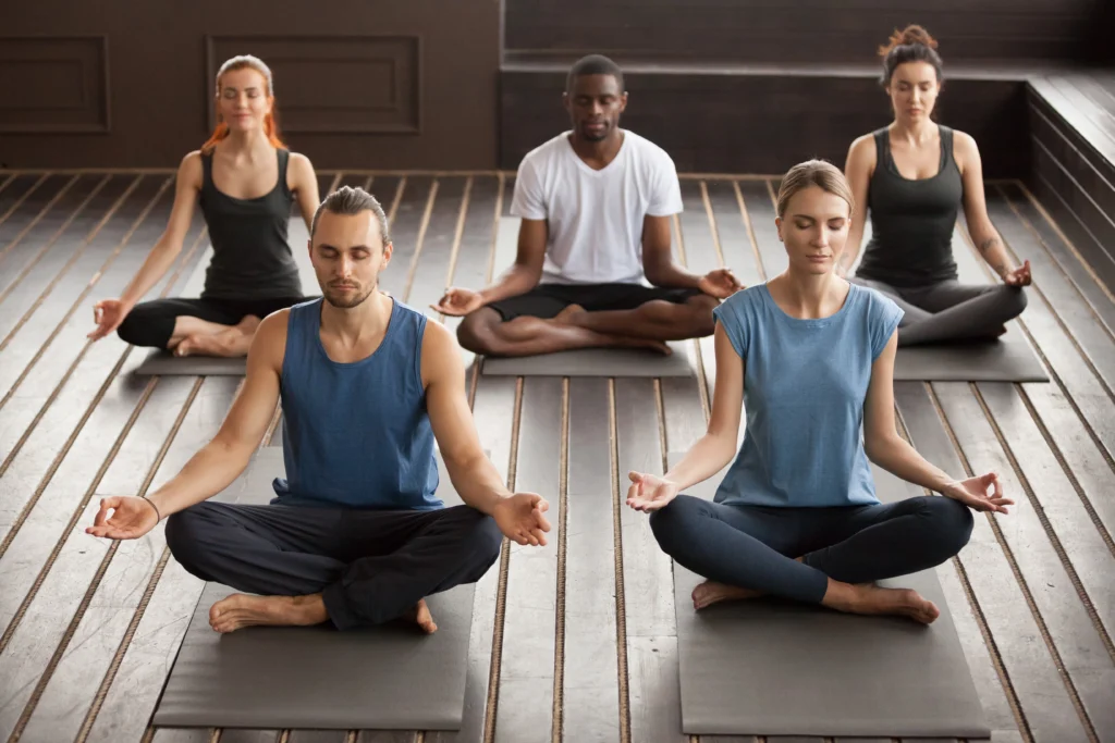 Group of people practicing meditation during yoga classes, sitting cross-legged in a calm studio with peaceful focused expressions.