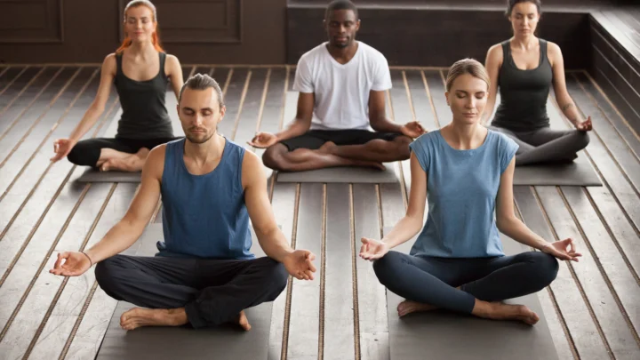 Group of people practicing meditation during yoga classes, sitting cross-legged in a calm studio with peaceful focused expressions.